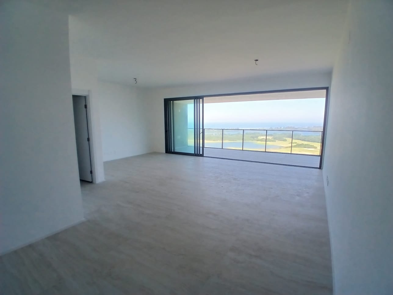 Sala ampla vazia da cobertura no Atlântico Golf, na Barra da Tijuca, com piso claro, porta de vidro de correr para a varanda e vista para o campo de golfe, a lagoa e o mar
