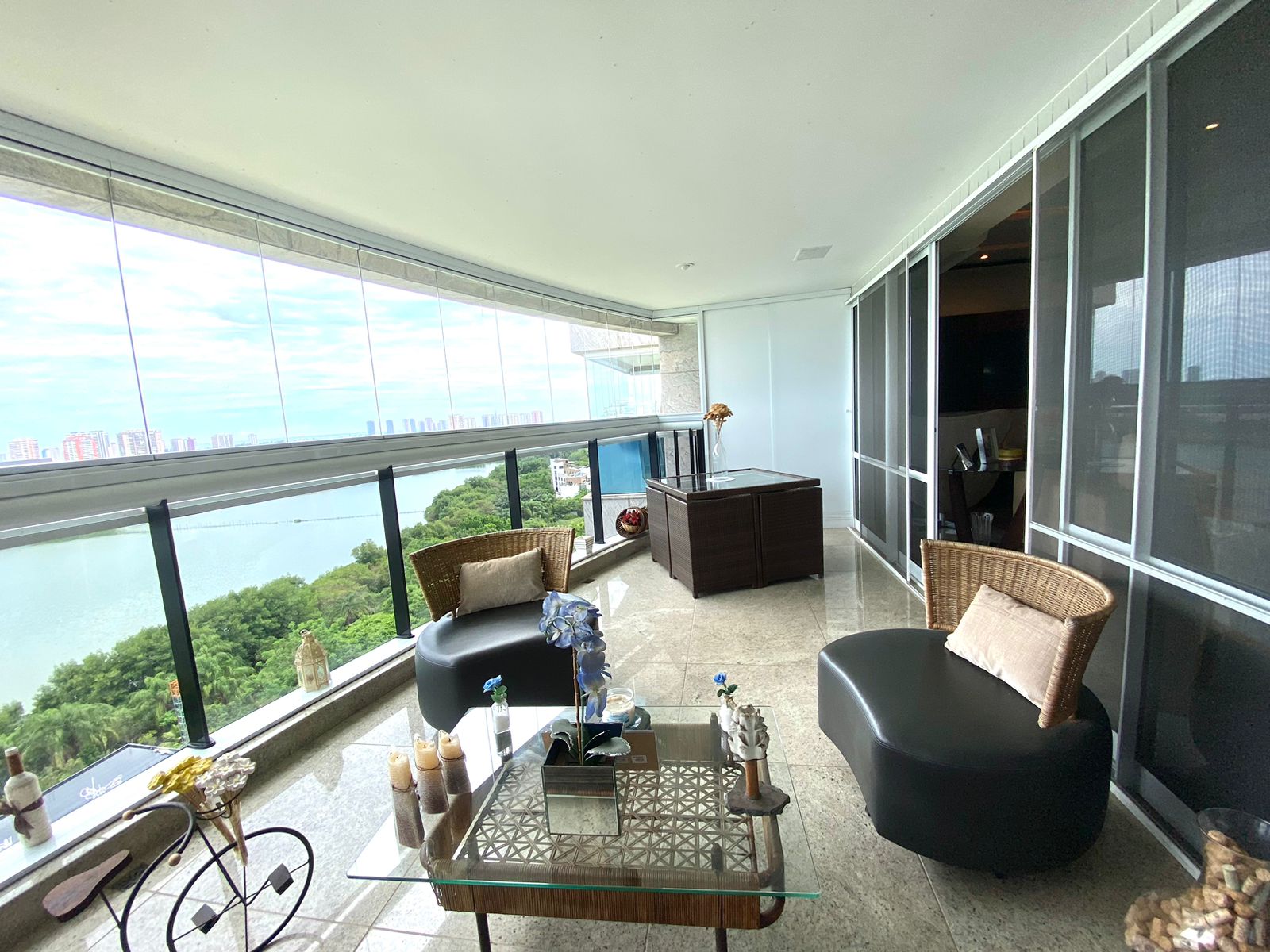 Varanda com cortina de vidro do apartamento na Península Aquarela, na Barra da Tijuca, com poltronas de vime, mesa de vidro e vista para a lagoa e a cidade