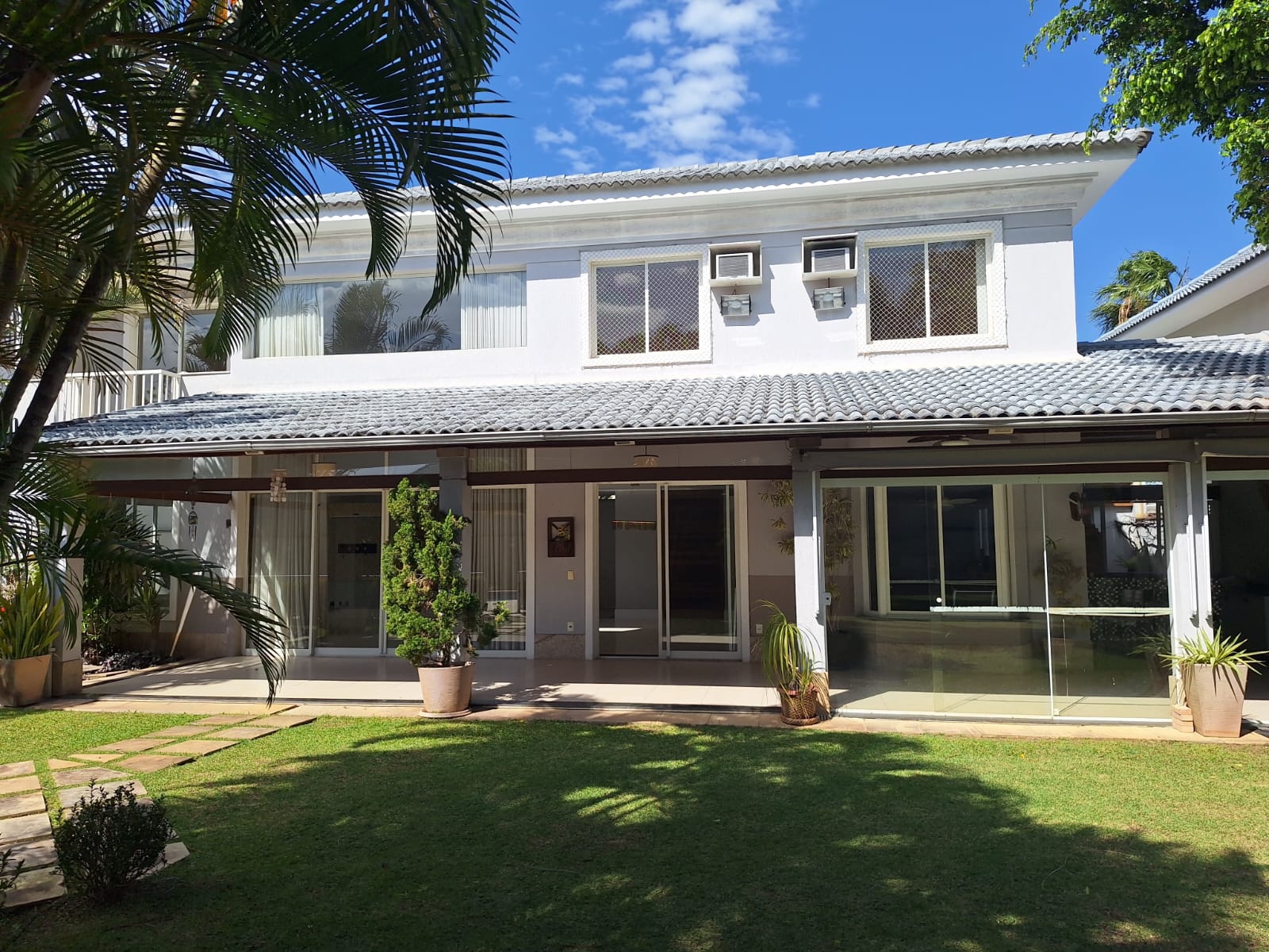 Fachada da casa no Santa Mônica Jardins, na Barra da Tijuca, com dois andares, telhado de telhas, jardim gramado e palmeiras