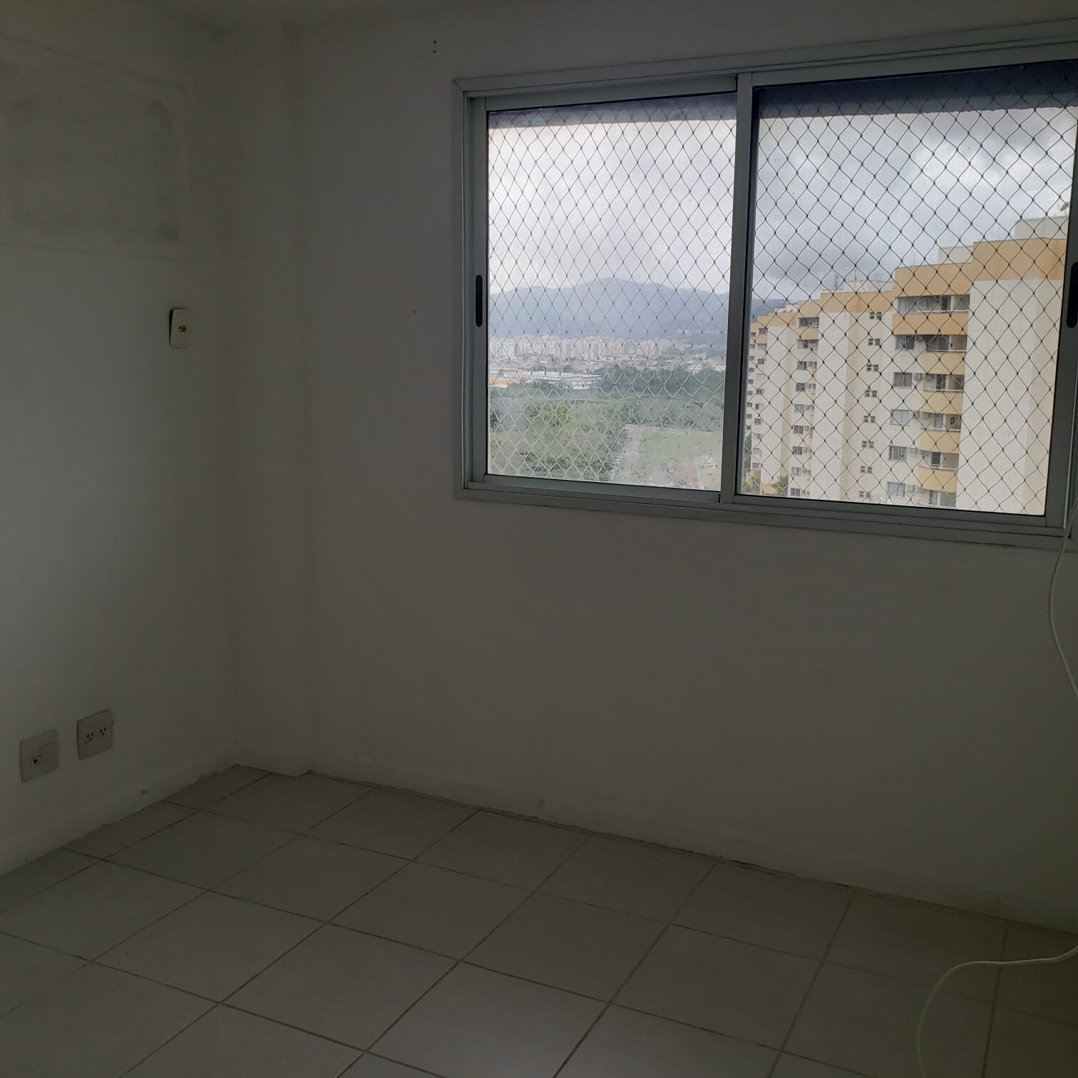 Quarto do apartamento na Vila do Pan, com janela e vista de prédios e morros
